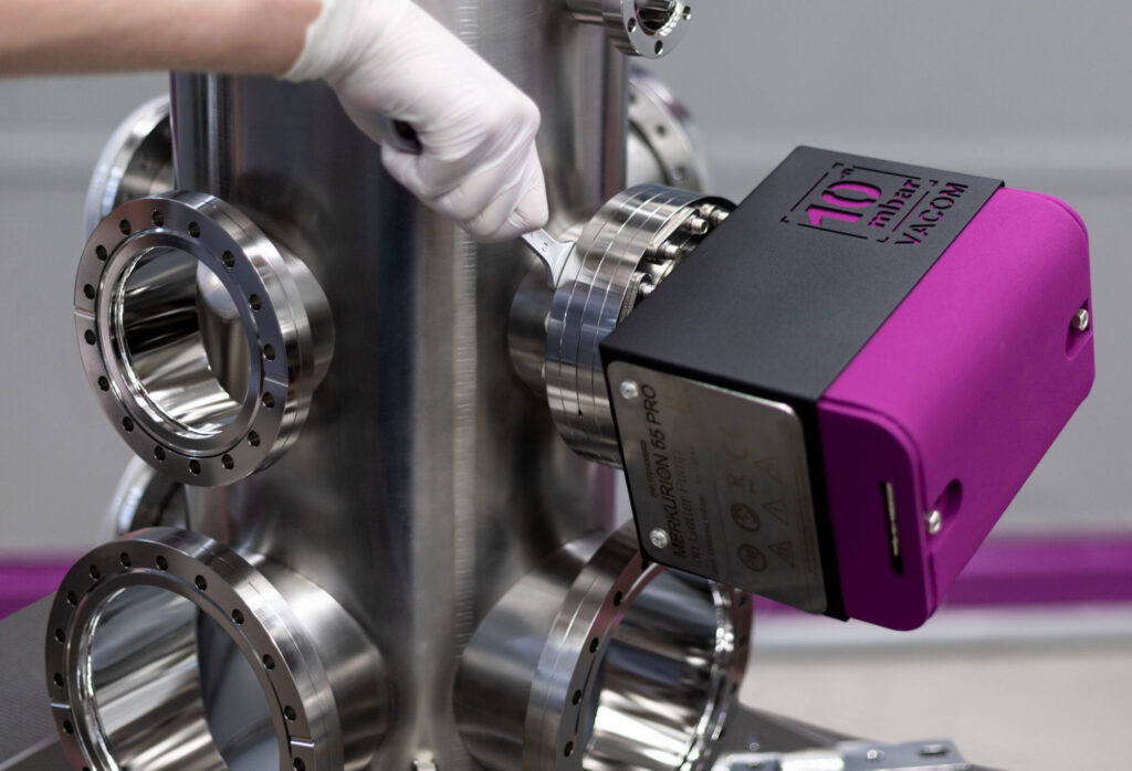 A product photography of an Ion getter pump named Merkurion by VACOM 2021. A hand montages it to a vacuum chamber.